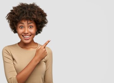 horizontal-shot-of-pretty-dark-skinned-woman-with-afro-hairstyle-has-broad-smile-white-teeth-shows-something-nice-to-friend-points-at-upper-right-corner-stands-against-wall